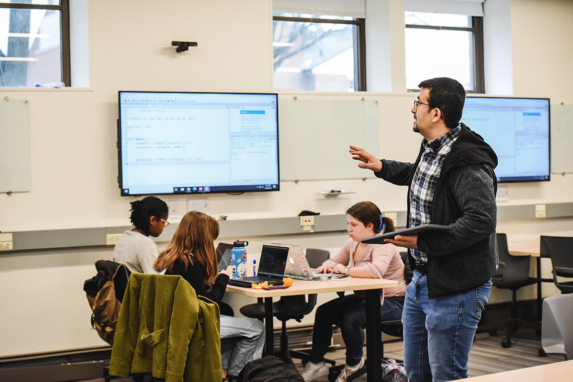 A college professor walks around the classroom as students at tables look at a large screen on the wall