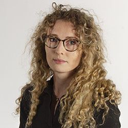 A headshot of Emily Potts, who wears glasses and has long, curly blonde/brown hair