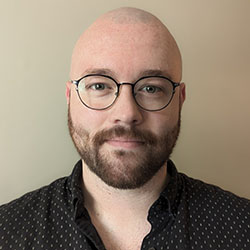 A headshot of Joseph Meadows, who is bald with dark facial hair and wears glasses