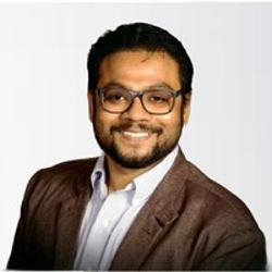 A headshot of Apoorva Ghosh, who has short dark hair and glasses and wears a brown blazer