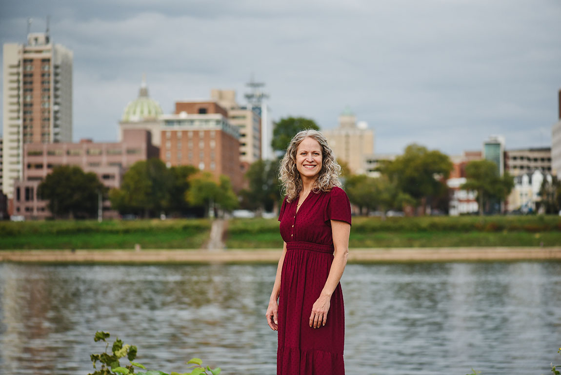 A woman with shoulder-length curly hair wearing a red dress smiles at the camera as she stands along the Harrisburg riverfront with the city skyline behind her