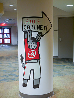 A cartoon mule wearing a red T-shirt and white shorts holding an arrow-shaped sign that says M.U.L.E. Cabinet! is painted on a column inside a building