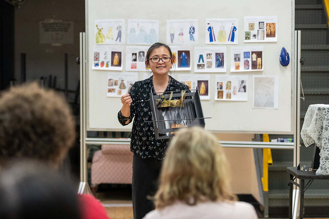 A professor with dark hair and glasses smiles as she presents a set model to a group of performers