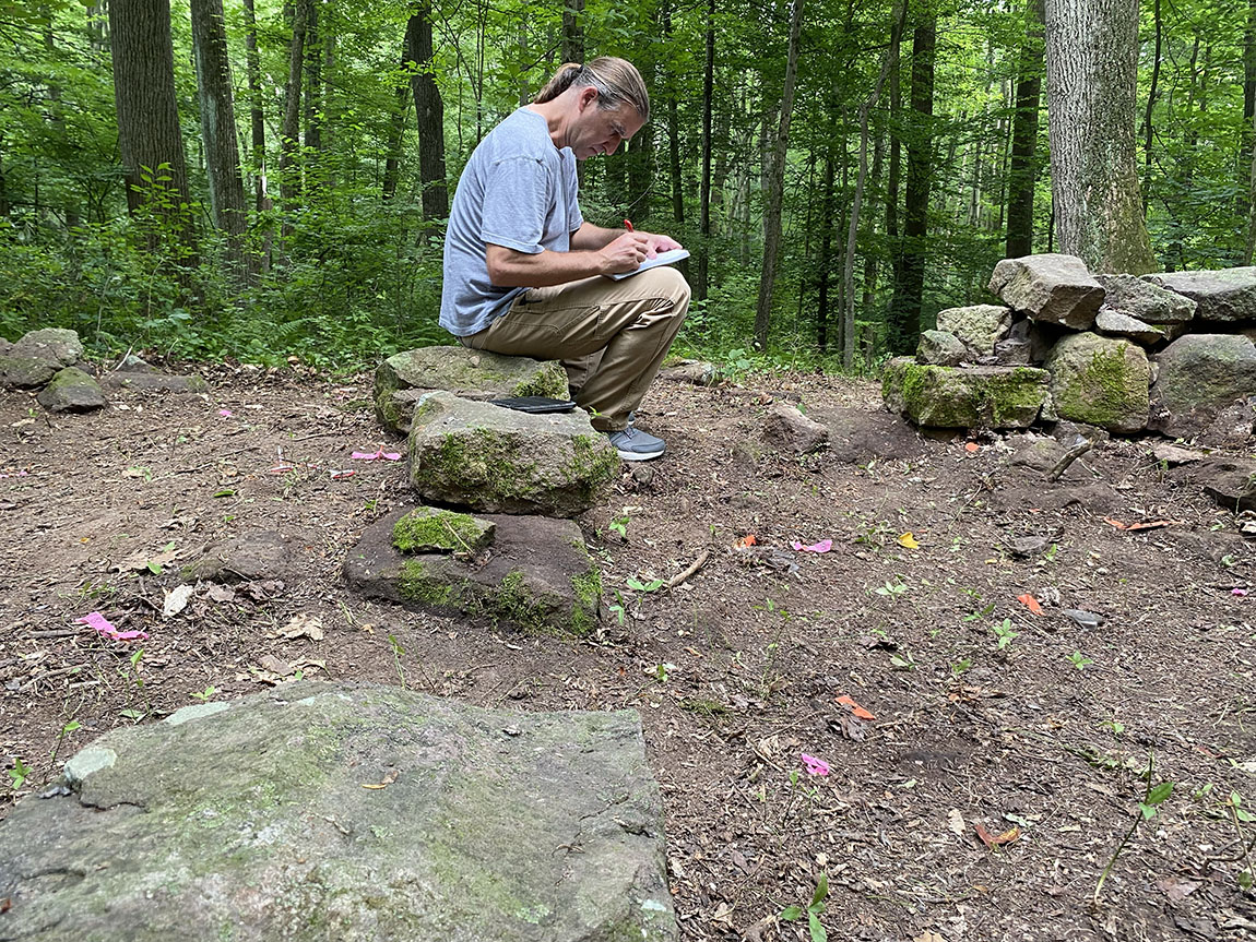 A college professor sits on some rocks in the woods and writes in a notebook