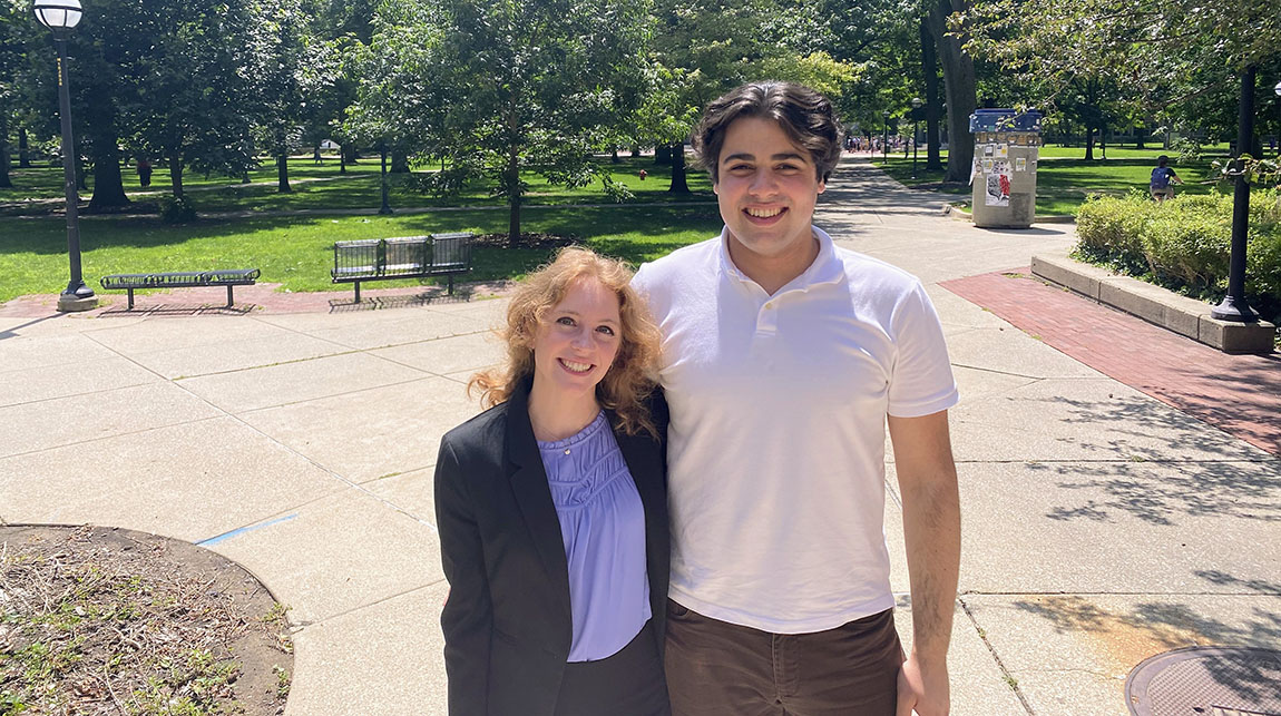 Two college students, one with long light hair and one with short dark hair, smile at the camera outside on a college campus