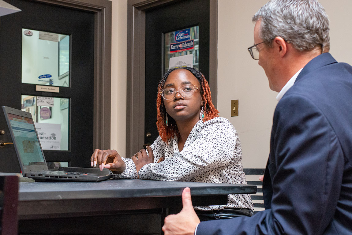 A college student with shoulder-length hair and glasses sits next to and listens to a college professor, with a laptop open in front of them