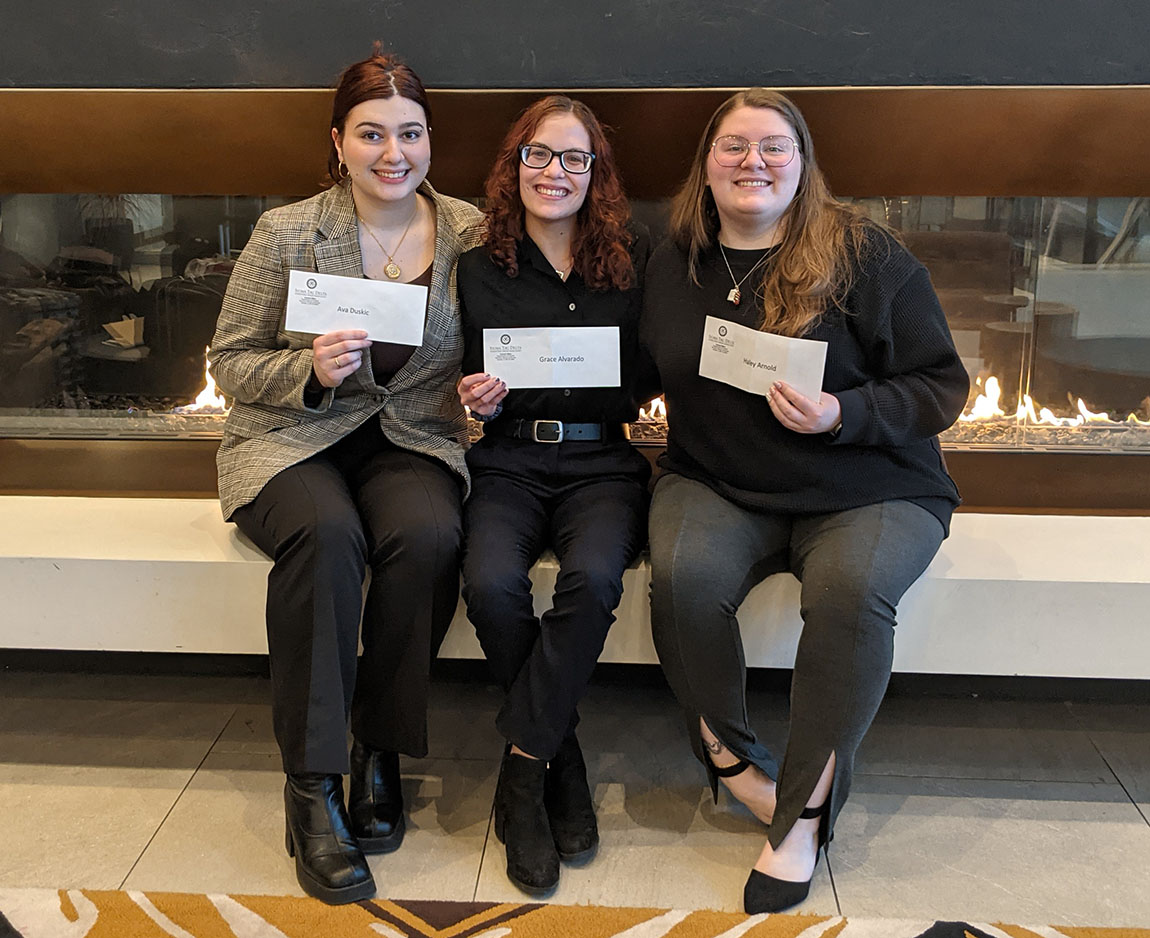 Three college students smile and sit in front of a fireplace holding up white envelopes containing a reward