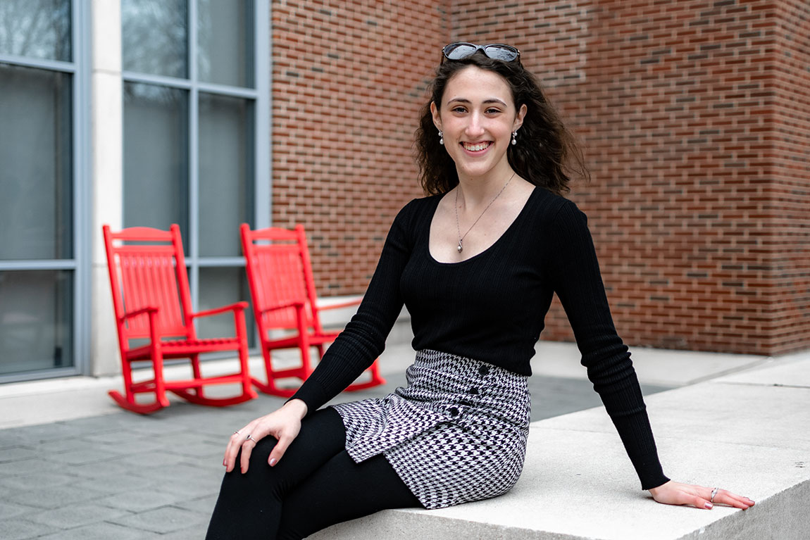 A college student in black shirt and black and white skirt sitting outside on a wall and smiling with two red rocking chairs in the background