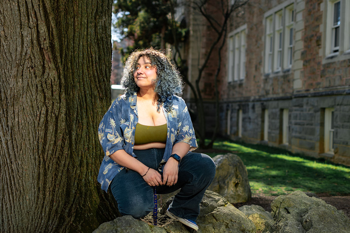 A college student with long curly hair sits beneath a tree and looks off into the distance