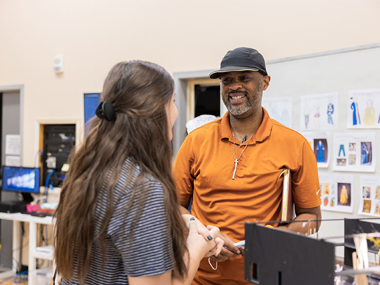 An instructor in a black ball cap and orange shirt speaks to a student in a studio space.