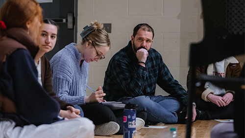 An instructor sits on the floor of a studio, head in hand, while listening to students nearby speak.