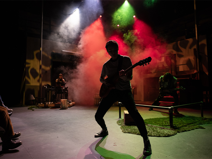 A person, seen in silhouette, plays guitar on a small stage with multicolor lights reflected in fog spilling from the ceiling.