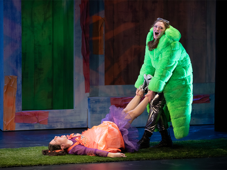 A student in a lime green fuzzy coat drags another students wearing an orange top and tutu, along the floor of a stage.