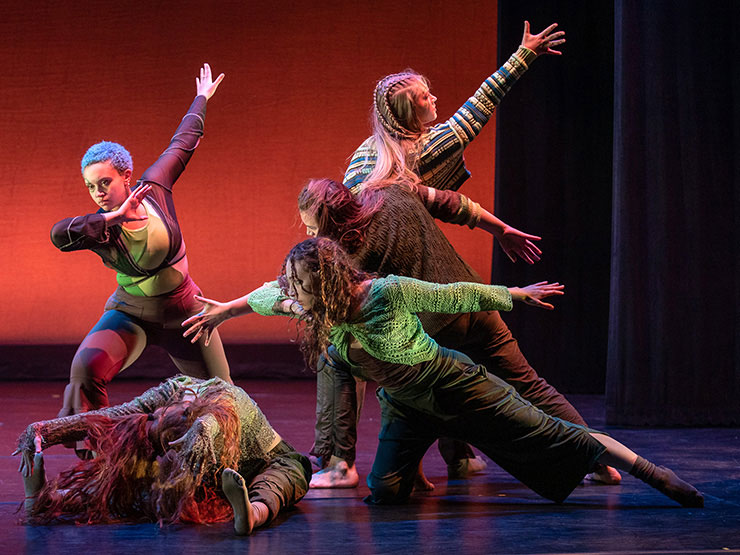 A group of dancers move dramatically on a stage with warm orange lighting.