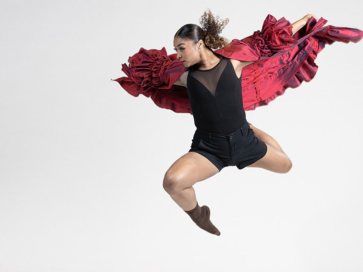 A young adult in a black dancing outfit leaps in the air with arms, draped with a ruffled red cape, outstrecthed.