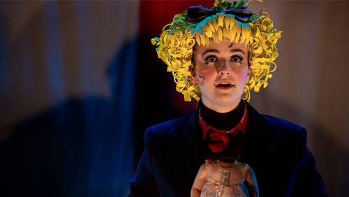 An expressive college student performs on stage dressed as a doll.