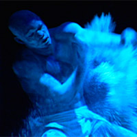 photo under blue light of an African dancer in traditional makeup. It appears that either his arms are motioned-blurred, or he is scattering a powder, or both.