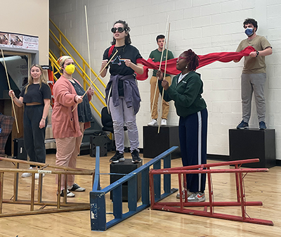 Six college-age actors work in a rehearsal hall. Several ladders lie on their sides on the floor, forming the shape of an airplane. One actor stands on a small box in the middle of the ladders, wearing aviator goggles and miming holding airplane controls. The other actors hold sticks and long, flowy fabric, meant to simulate the appearance of an airplane flying.