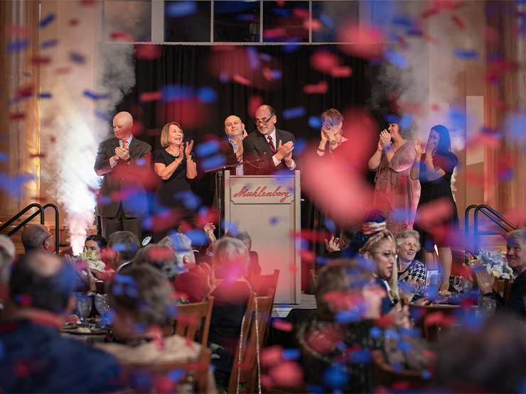 A group of people clap on stage as blue and red confetti rains down from above.