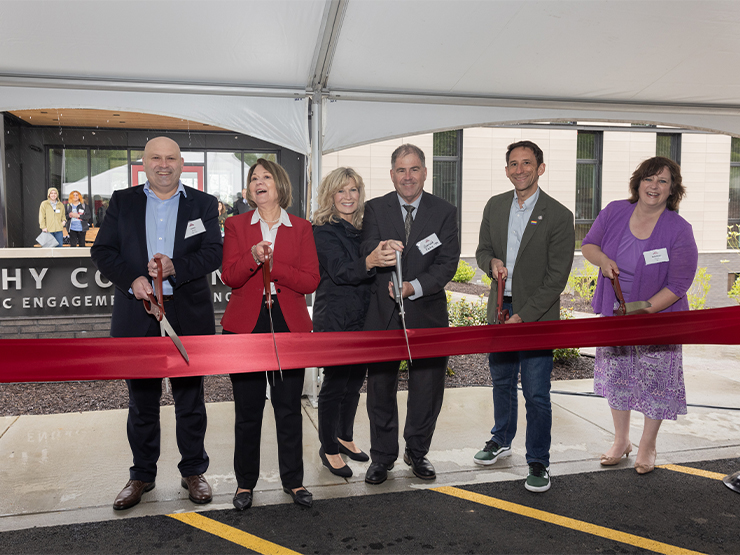 A group of men and women, dressed formally, stand under a tent and pose with oversized scissors, prepared to cut a red ribbon strung in front of them.