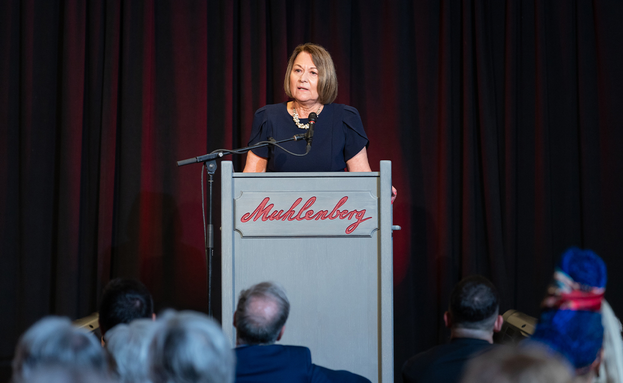 A woman in a navy blue dress speaks to a crowd behind a podium that reads 