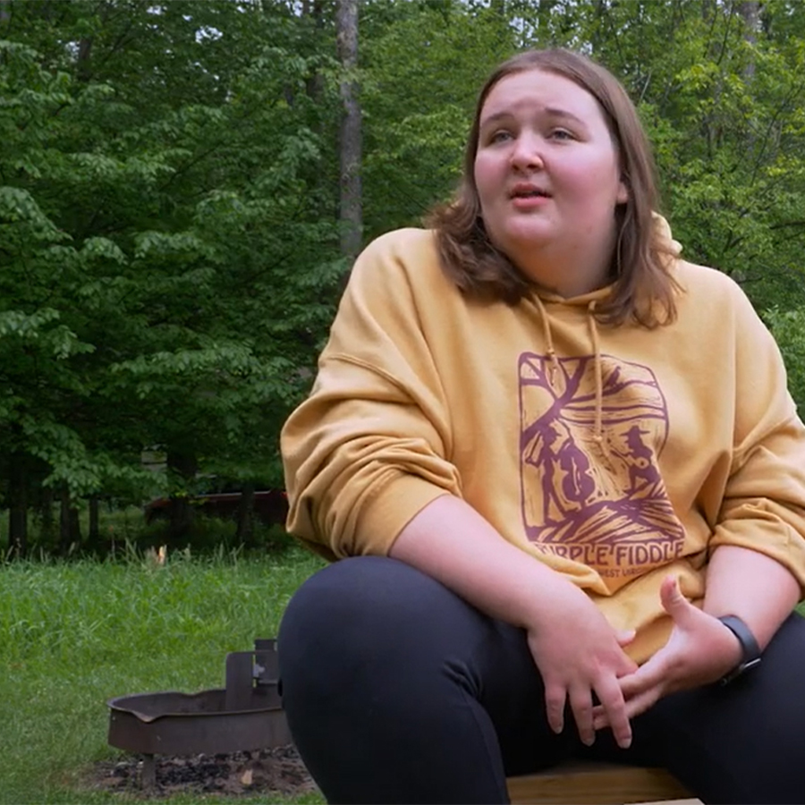 A students in a yellow sweatshirt speaks to the camera while sitting on a picnic table at a campground surrounded by woods.