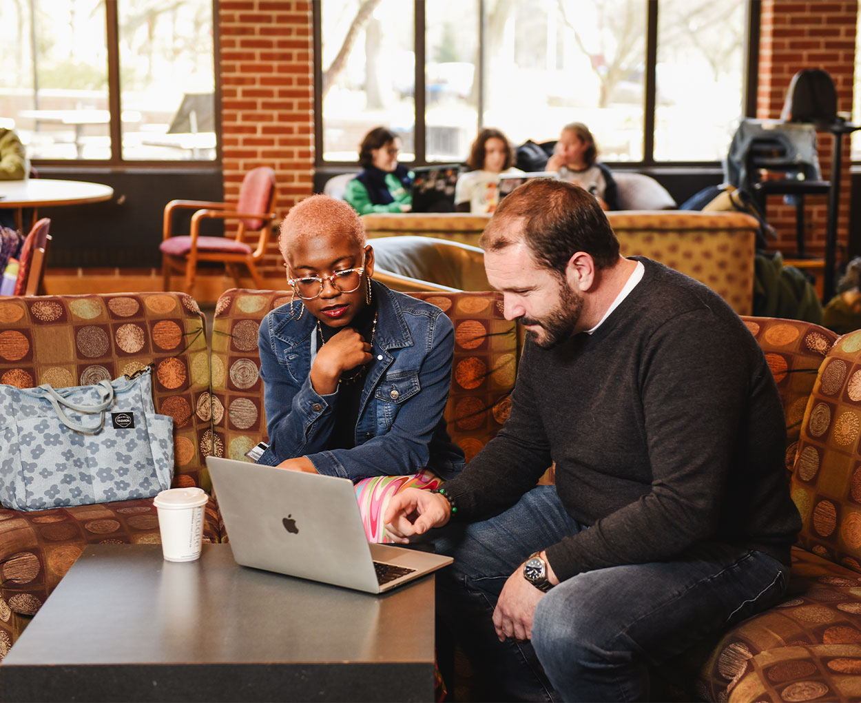 A student sits near an instructor on a couch at a college coffee shop as they review content on an open laptop between them.