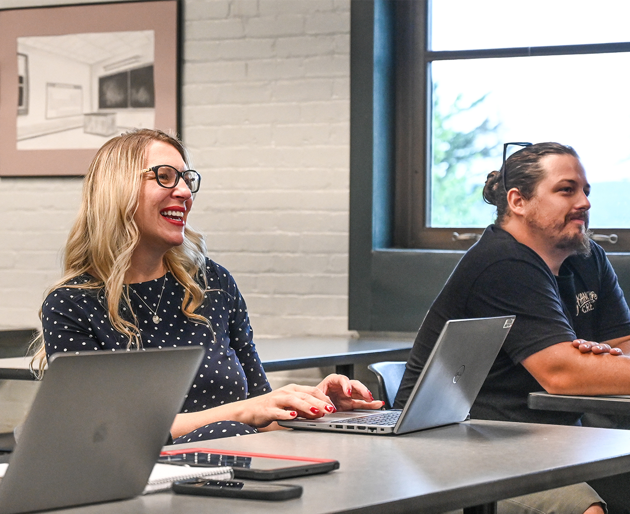 Two adults smile while sitting at tables in a classroom, with open laptops in front of them.