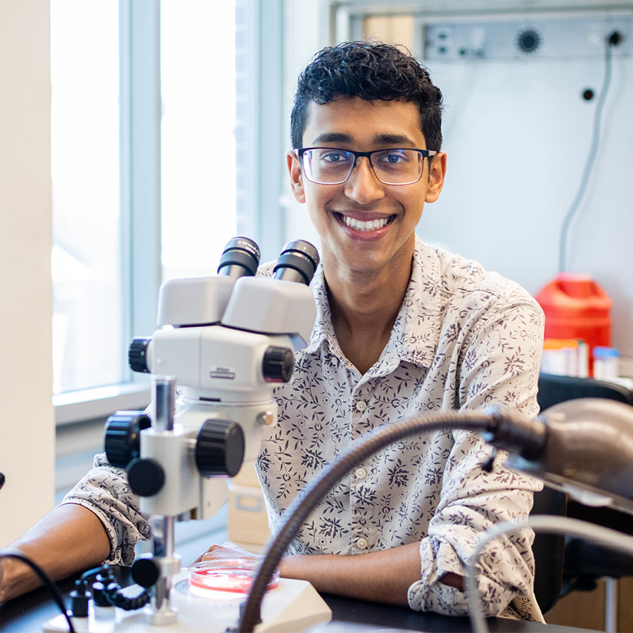 A young adult in a checkered shirts smiles at the camera from behind a microscope in a science lab.