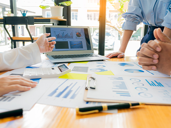 A group of people gather around a table scattered with documents, spreadsheets and a laptop.