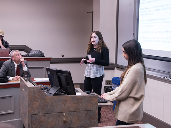 Two students stand in front of a classroom, giving a presentation, while a man in a suit looks on, chin resting in his hands.