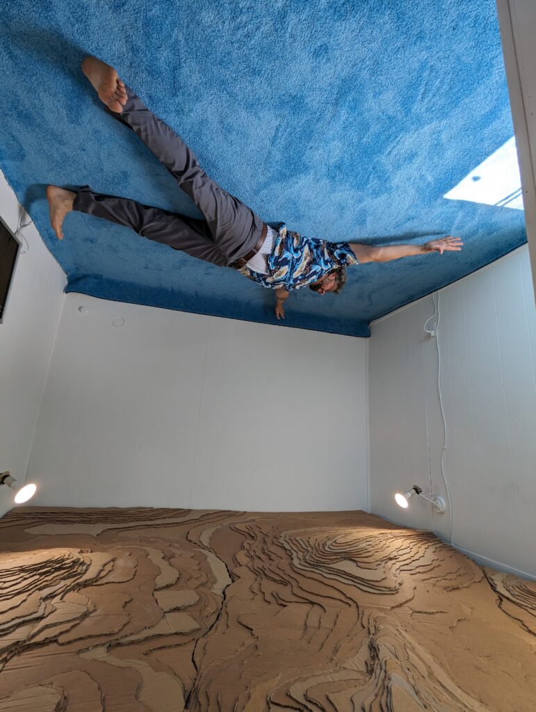 A man lies on a blue carpet above a cardboard relief map, seemingly defining gravity.