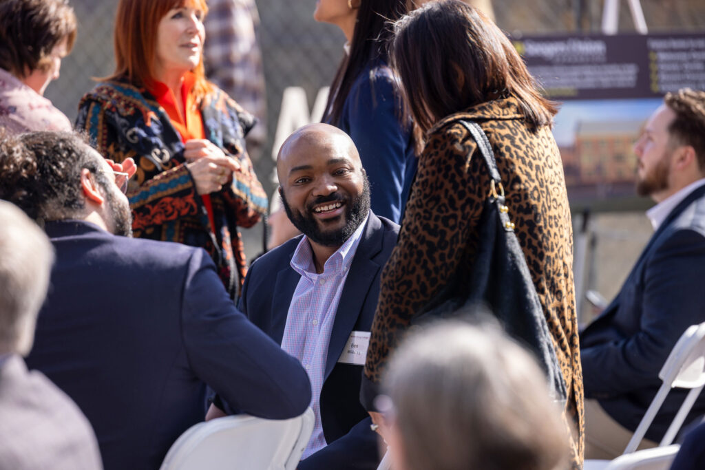 A man with a beard sits and talks to a standing woman at an outdoor ceremony