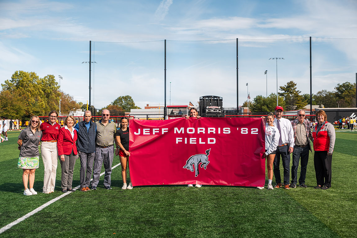 A group of people poses on a soccer field with a banner that says Jeff Morris '82 Field