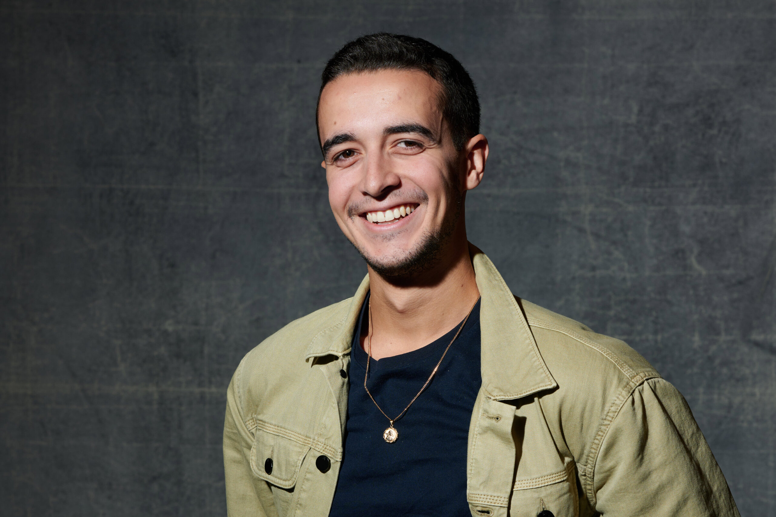 A man with dark hair in a green jacket smiles in front of a gray background