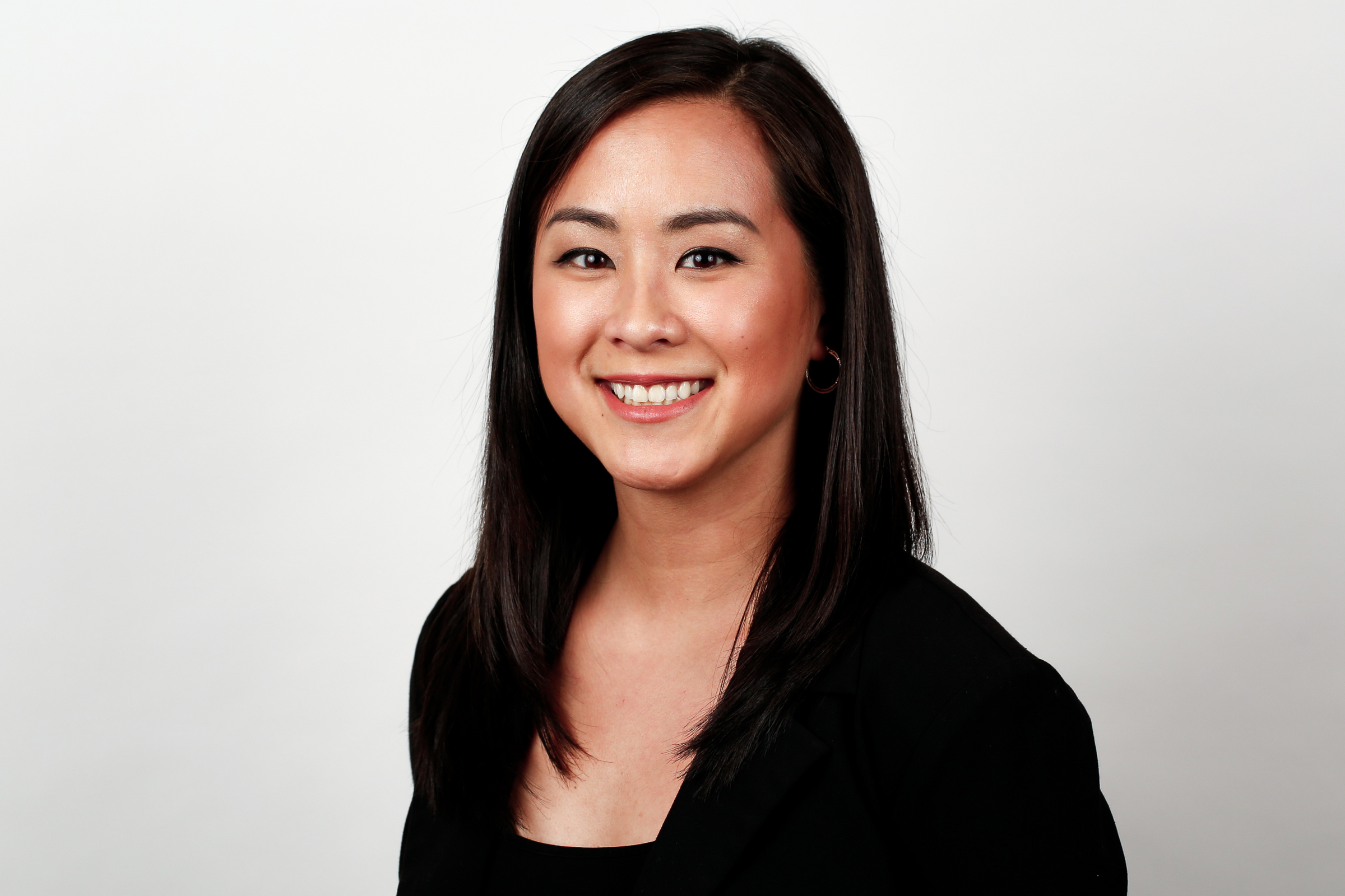 A woman with long dark hair in a black blazer smiles in front of a white background