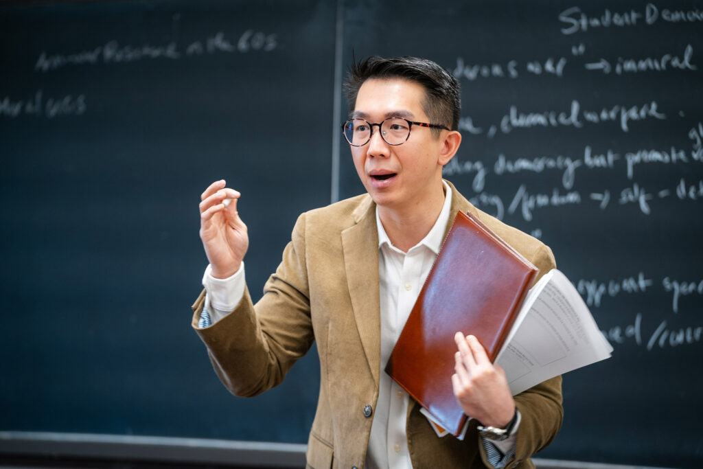 A college professor in glasses and a tan blazer teaches a class