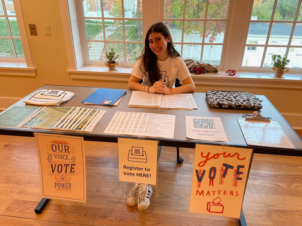 A college student sits at a table with signage that says "BERG VOTES Register to Vote HERE!"