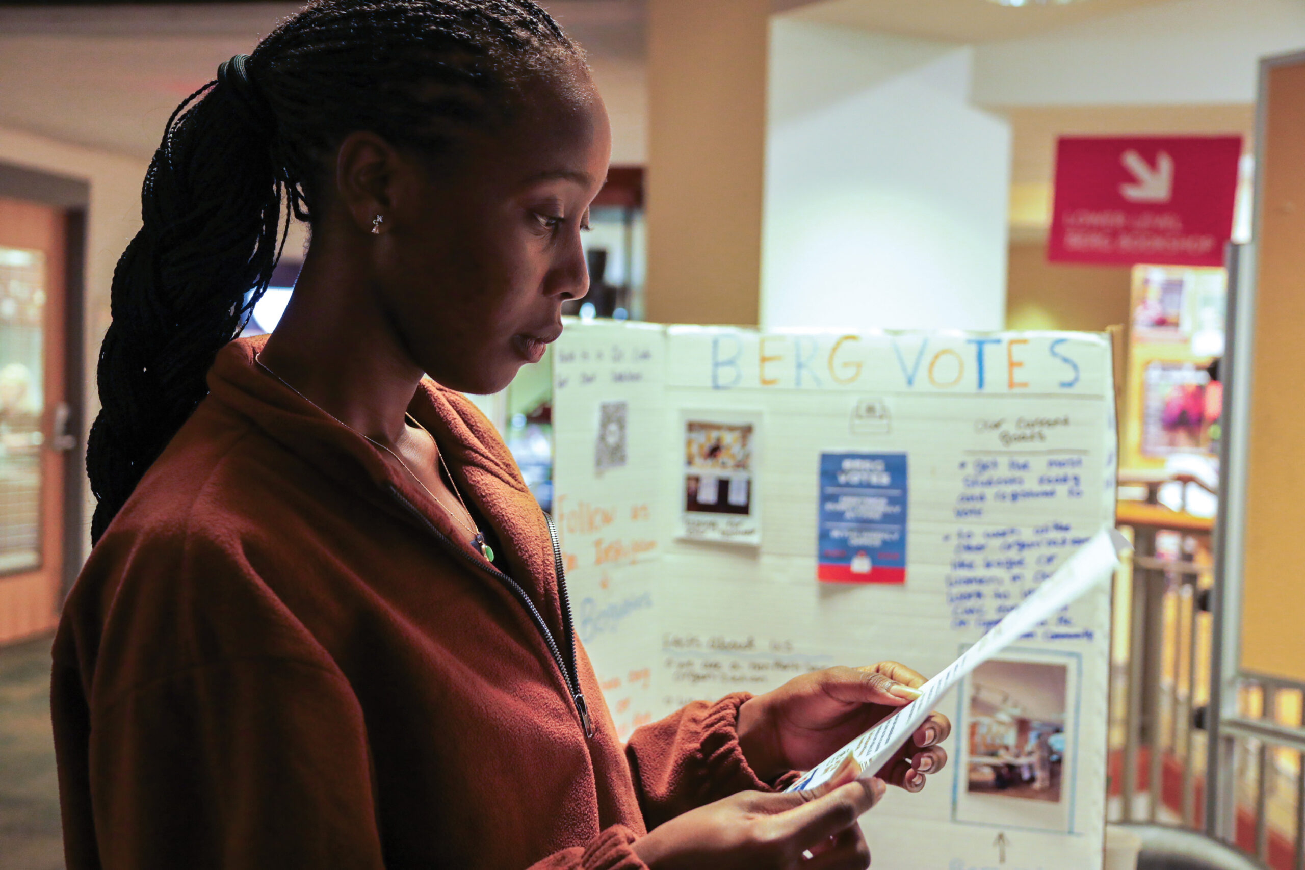 A college student reads a piece of paper in front of a sign that says Berg Votes