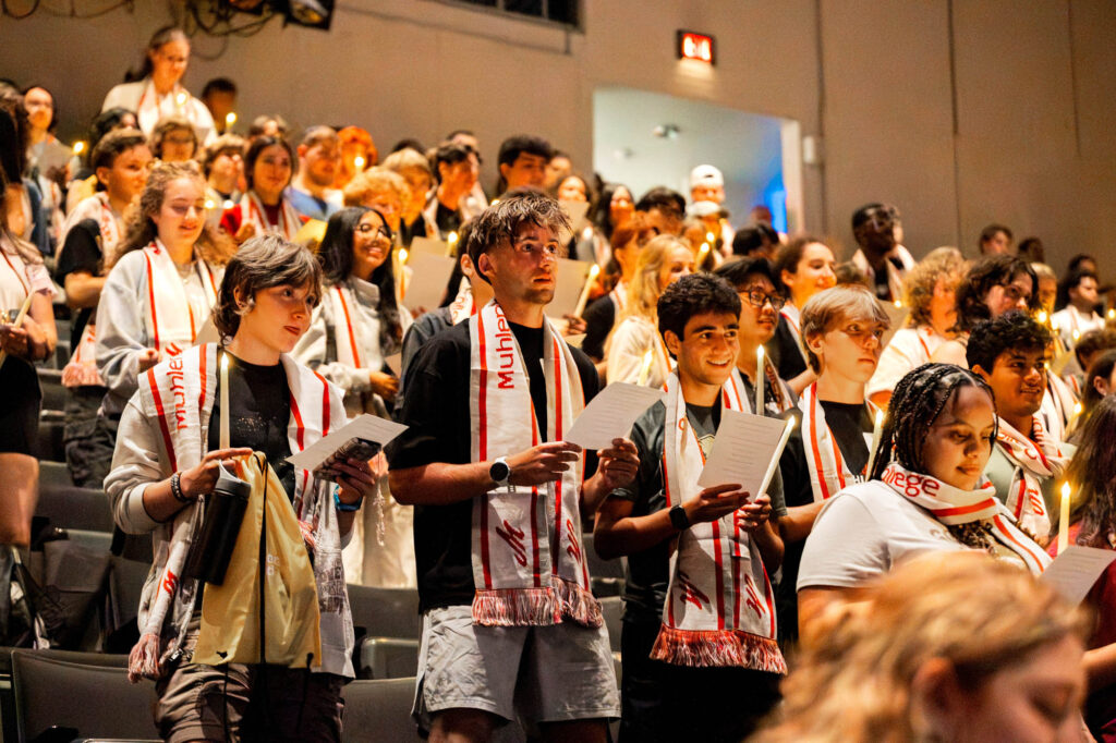 New Muhlenberg College students sing in an auditorium while wearing scarves and holding candles