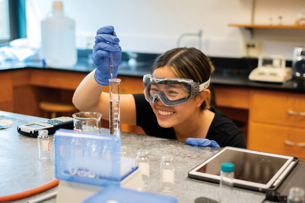 A Muhlenberg student wearing goggles adds liquid to a test tube in a lab.