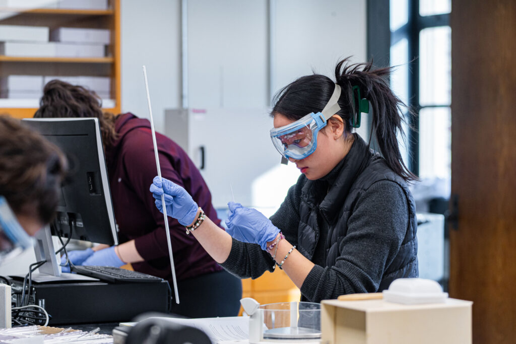 A Muhlenberg student with googles and gloves works in a chemistry lab.
