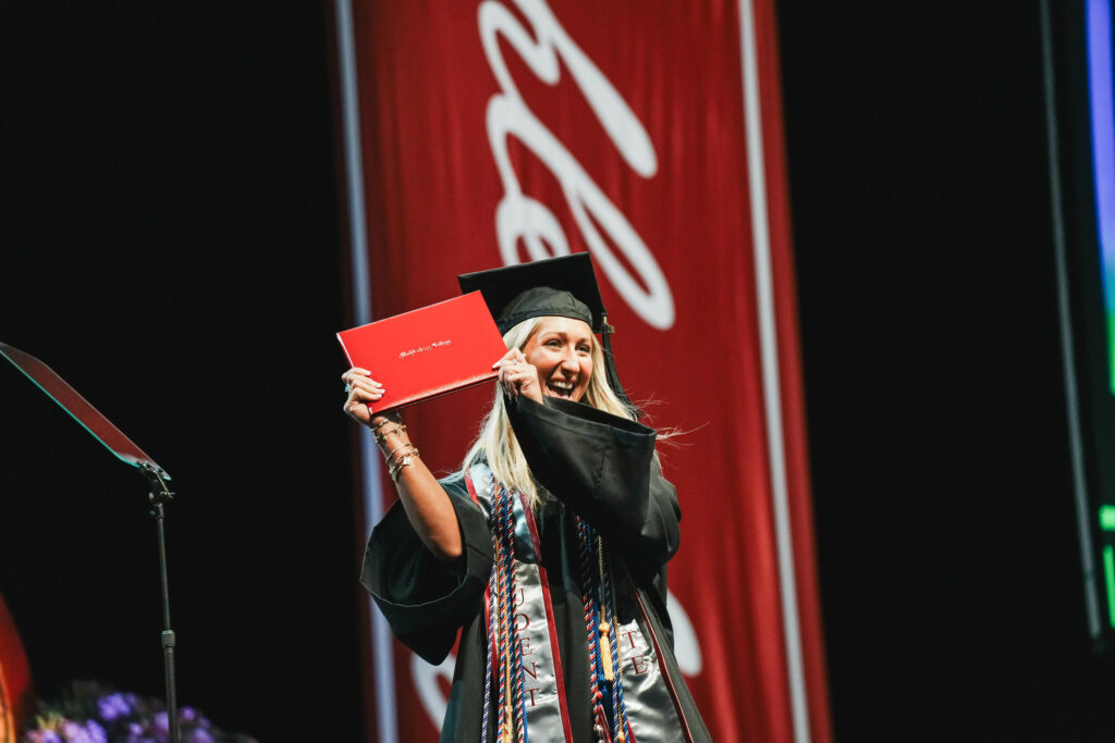 A college gradaute walks the stage at commencement and holds up her diploma