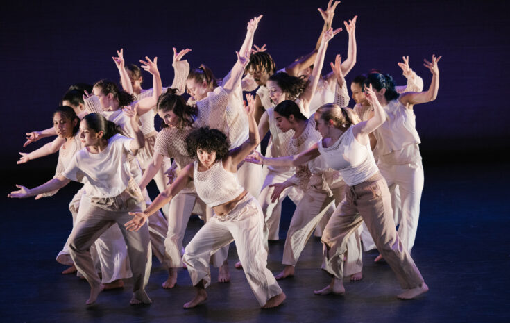 A group of college students in white shirts and tan pants do a choreographed dance on stage