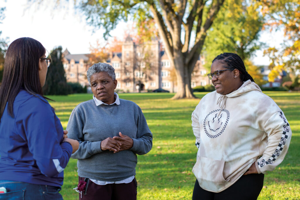 A college staff member talks to two college students on a grassy lawn