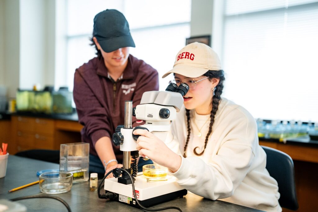Two students in a lab wearing Muhlenberg gear. One sits and looks into a microscope as the other talks a d watches.