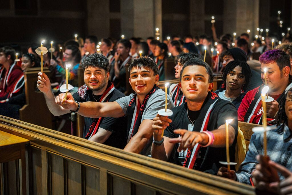 First-year college students sit in a chapel pew holding lit candles and wearing Muhlenberg College scarves