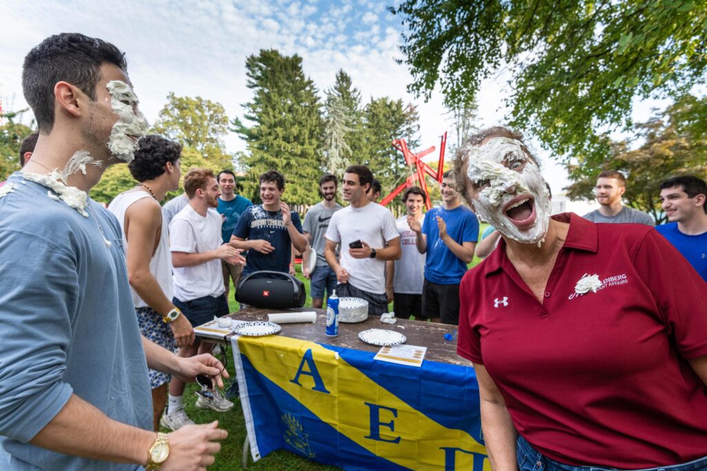 A college staffer has pie on her face next to a college student in front of a table with Greek letters and a group of fraternity members