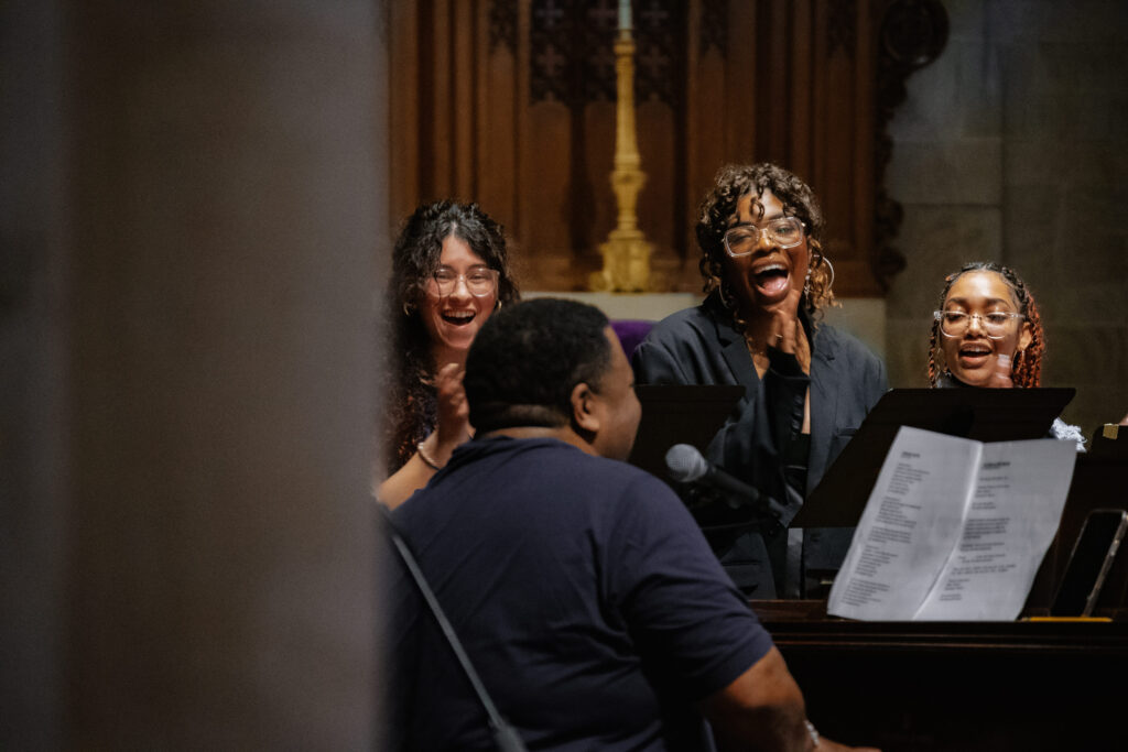 Three college students sing behind a piano a man is playing