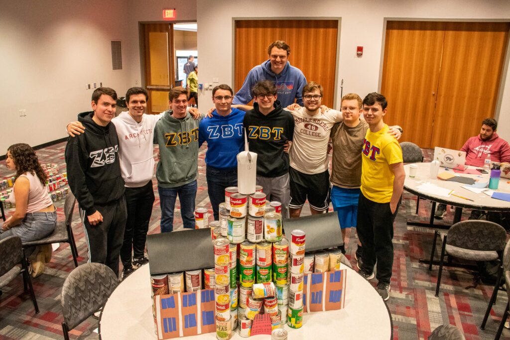 A group of fraternity members stand behind a tower of cans